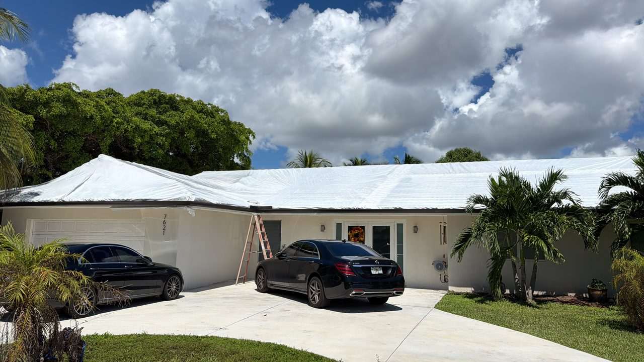 Ground-level view of a long single-storey home with white shrink wrap sealed across the whole roof, ladder still against the wall