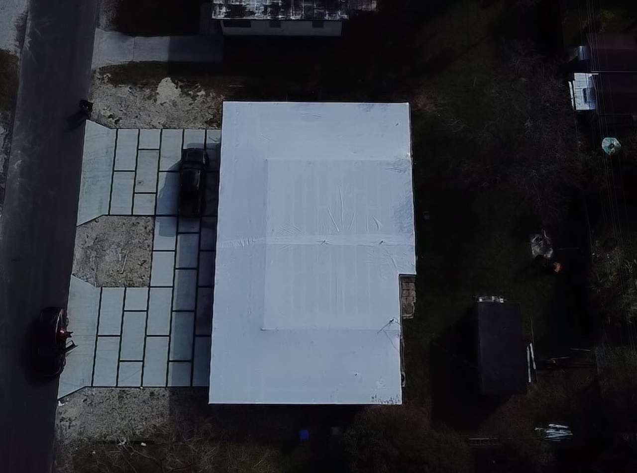 Overhead view of a rectangular roof fully sealed in white wrap, paved yard alongside