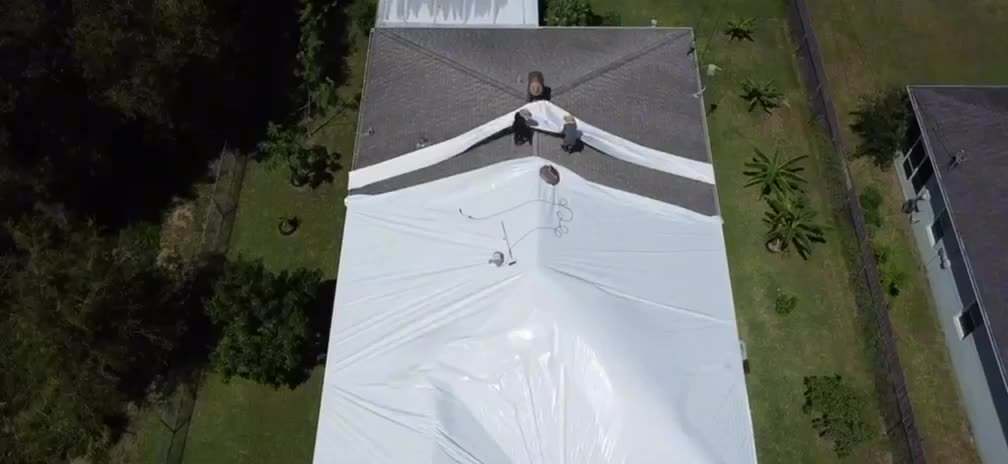 Aerial view of a Full House Wrap crew part-way through a job: half the roof still bare shingle, the other half already sealed under white shrink wrap
